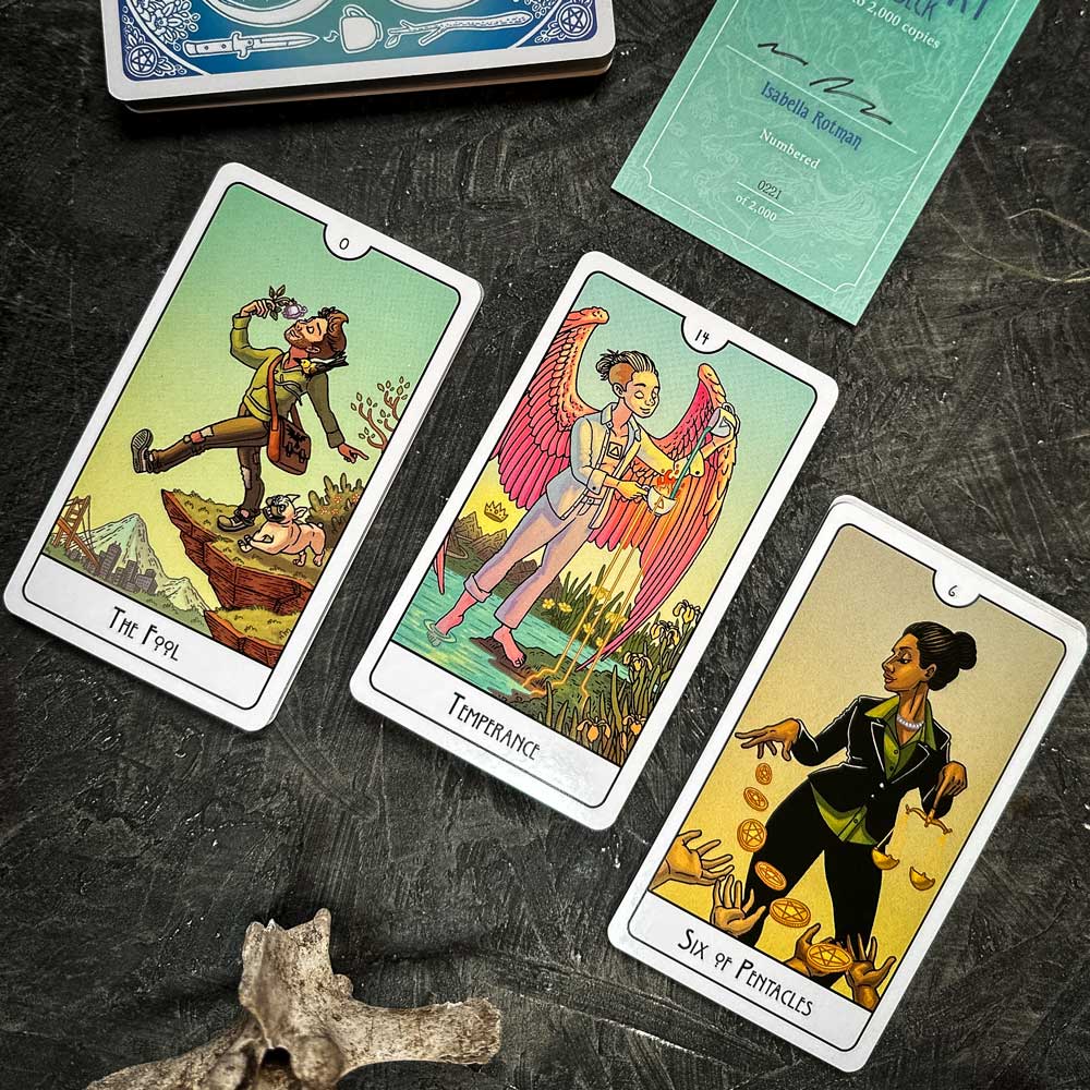A photo showcases The Fool, Temperance, and Six of Pentacles from the This Might Hurt Tarot - LE Boxed Set on a dark surface. Their vibrant illustrations are accompanied by a booklet and a small bone, enhancing the mystical ambiance.