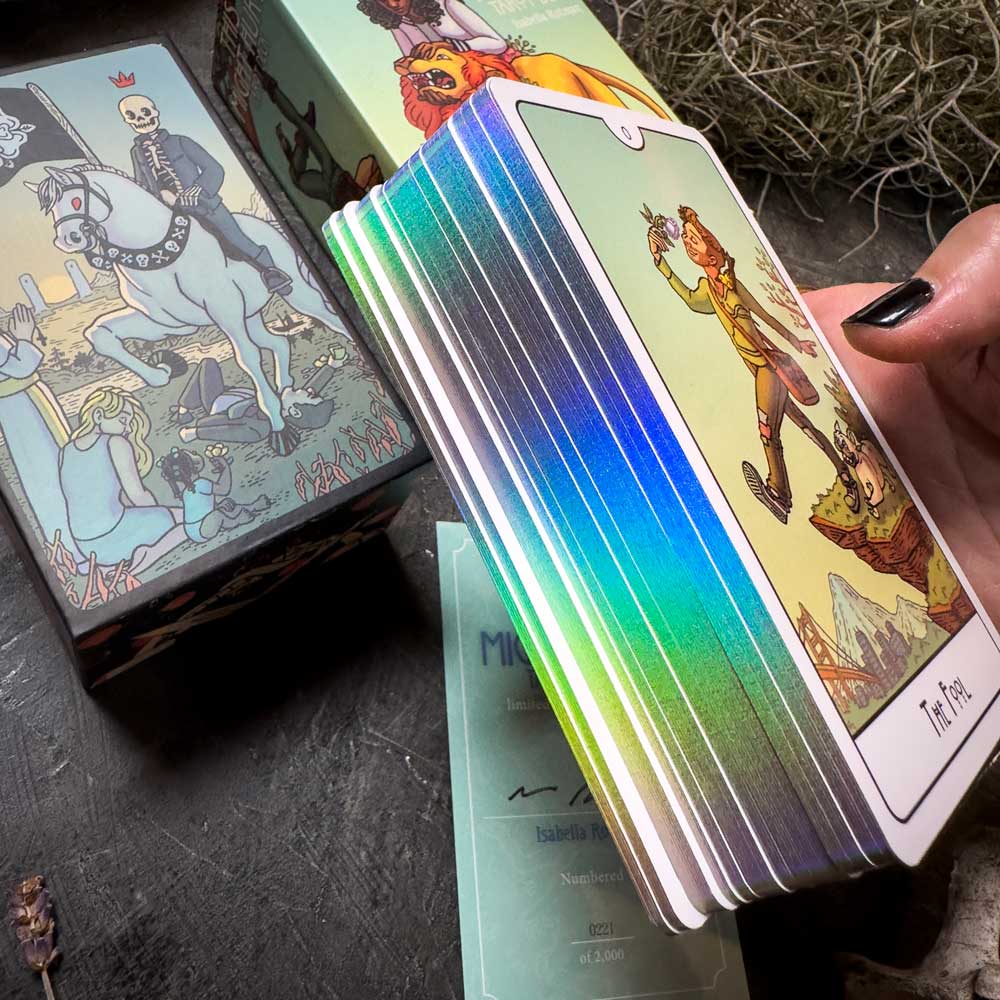 A hand holds the vibrant "This Might Hurt Tarot - LE Boxed Set" with "The Fool" card on top. The deck's iridescent edges shimmer while the box with a skeleton rider rests below on a dark, moss-covered surface, accompanied by a certificate.