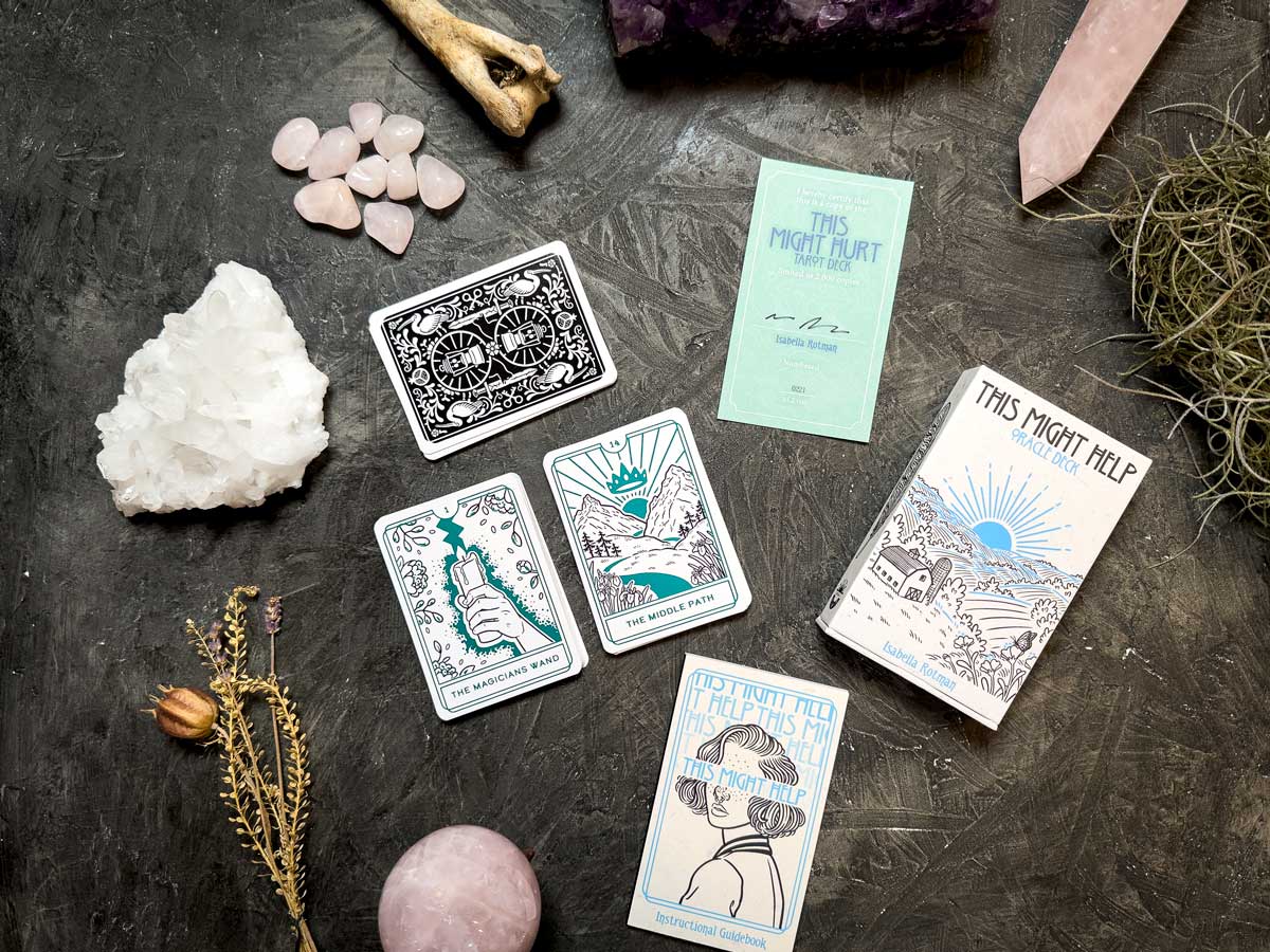 A set from the "This Might Hurt Tarot - LE Boxed Set" is displayed on a dark surface with crystals like rose quartz and amethyst, alongside a bone, dried plants, and a booklet labeled "This Might Hurt.