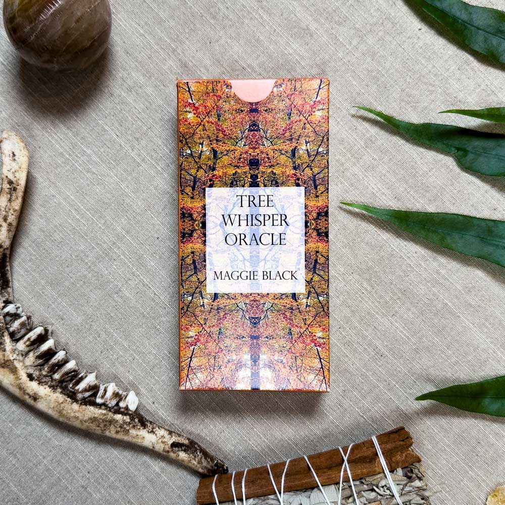 The "Tree Whisper Oracle" cards sit on fabric, surrounded by natural elements: a leaf, driftwood with teeth, and a mysterious brown object. The packaging displays an intricate tree design in rich autumn colors, capturing the essence of the oracle deck.