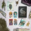 A flat lay of tarot cards, a black box with "The Unfolding Path Tarot" in silver, surrounded by crystals, moss, and a small tree branch. The cards depict diverse illustrations of people and nature, crafting an enchanting scene that suggests the journey ahead.