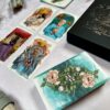A set of The Unfolding Path Tarot cards is spread out, with four intricately illustrated cards standing out. The card backs feature a floral design, and a decorative box nearby enhances the softly lit, mystical ambiance.