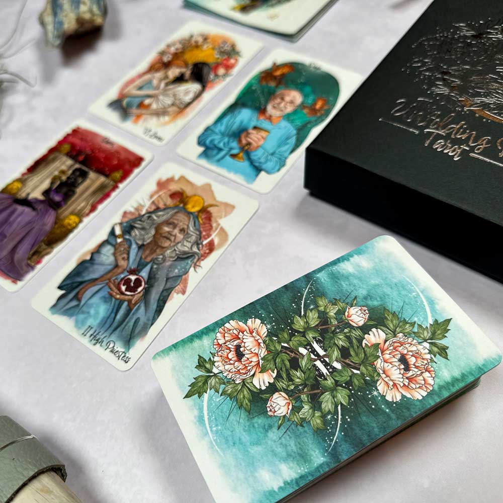 A set of The Unfolding Path Tarot cards is spread out, with four intricately illustrated cards standing out. The card backs feature a floral design, and a decorative box nearby enhances the softly lit, mystical ambiance.