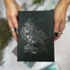 Hands with black nail polish hold the product "The Unfolding Path Tarot," featuring a box with intricate, shimmering silver floral artwork.