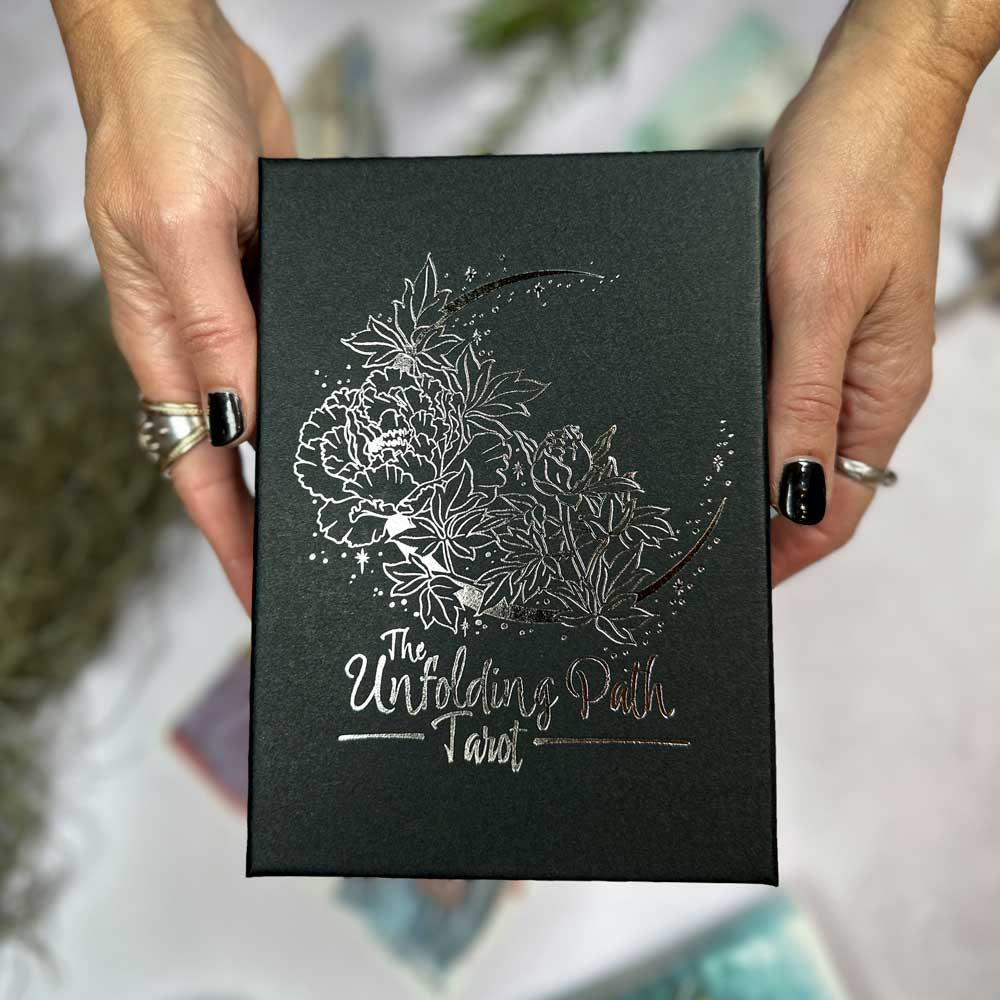 Hands with black nail polish hold the product "The Unfolding Path Tarot," featuring a box with intricate, shimmering silver floral artwork.