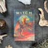 The book "W.I.T.C.H." lies on a dark, textured surface, its cover showing a witch riding a dragon. Natural elements such as stones and twigs are scattered around the book.