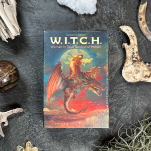 The book "W.I.T.C.H." lies on a dark, textured surface, its cover showing a witch riding a dragon. Natural elements such as stones and twigs are scattered around the book.