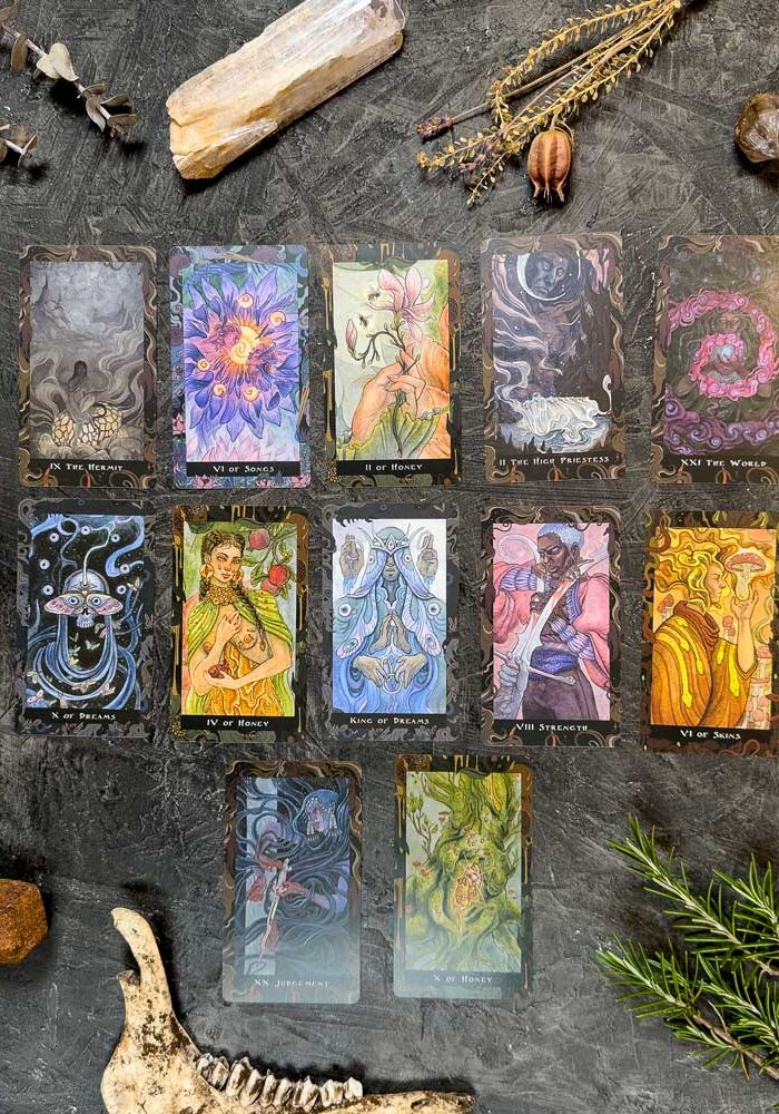 A set of 12 vibrant Blood Moon Tarot - Deluxe cards is elegantly displayed on a textured gray surface, surrounded by stones, herbs, a bone, and dried plants to create a mystical and earthy atmosphere.