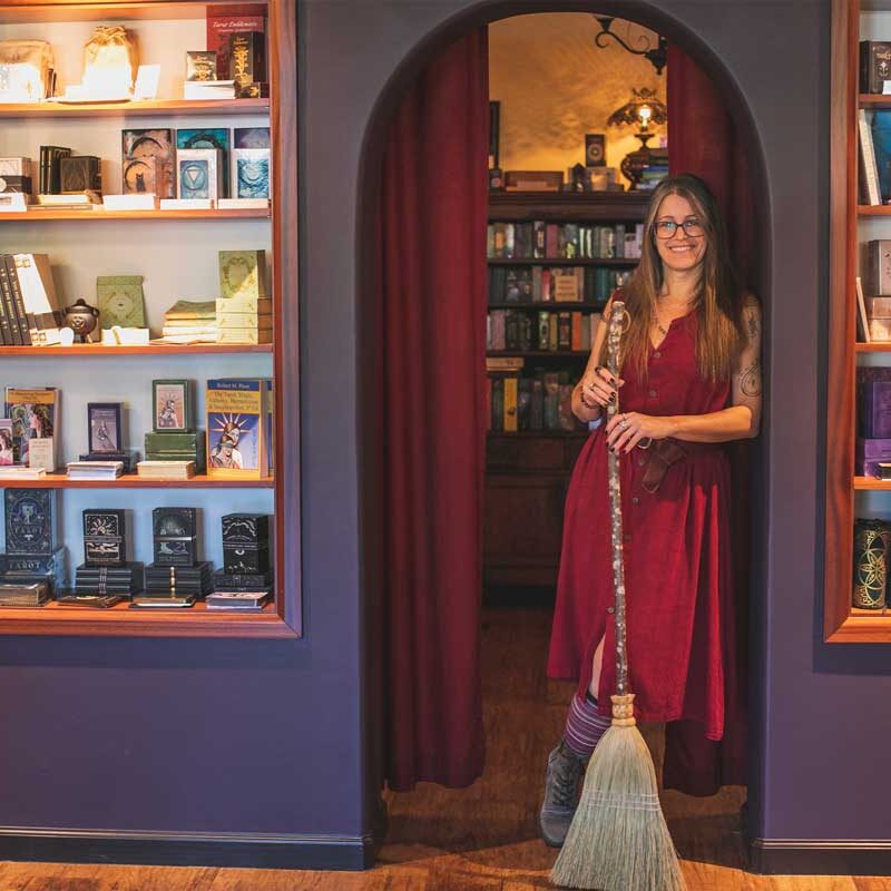 A woman in a red dress stands holding a broom in a cozy divination shop. Shelves filled with mystical books and items line the walls, evoking the metaphysical. A curtain behind her reveals more books and a warm, inviting atmosphere.